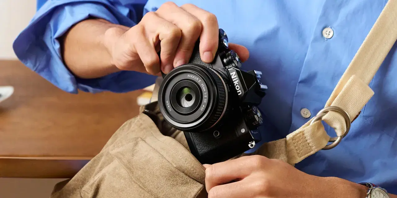 Osoba w niebieskiej koszuli wkłada czarny aparat Nikon z obiektywem do torby.