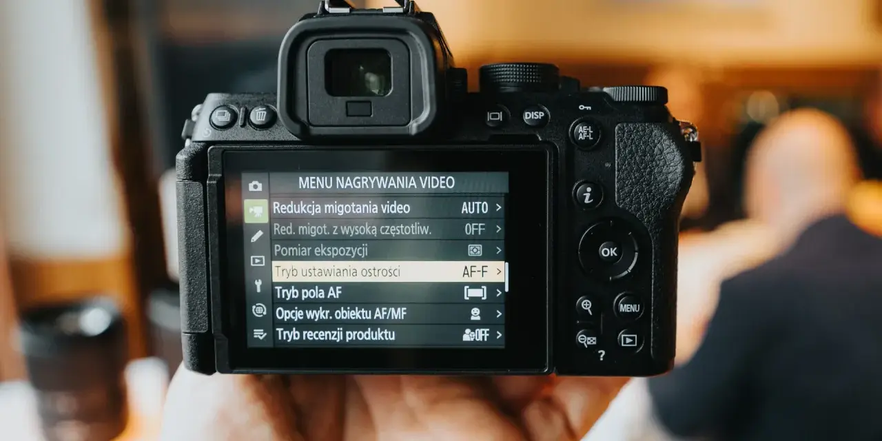 Dłoń trzyma aparat Nikon z włączonym menu nagrywania wideo, widoczne są opcje ustawień.