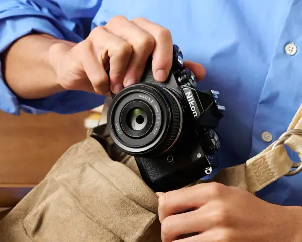 Osoba w niebieskiej koszuli wkłada czarny aparat Nikon z obiektywem do torby.