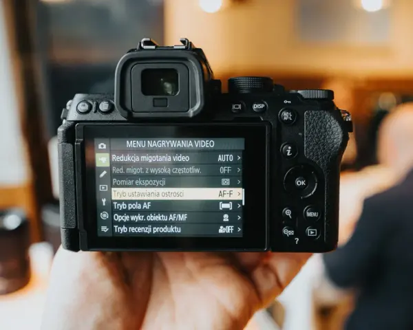 Dłoń trzyma aparat Nikon z włączonym menu nagrywania wideo, widoczne są opcje ustawień.